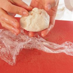 手順8：6で使ったラップに生地をスプーンですくって広げる。約大さじ1の餡をのせ、ラップで茶巾絞りのように包む（具は多少余る）。ベーキングシートの上にのせる。